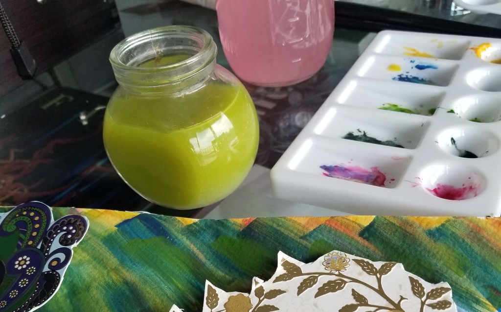 View of jars with colored water and a paint palette. In the foreground is a canvas with various colors and some paper cut outs.