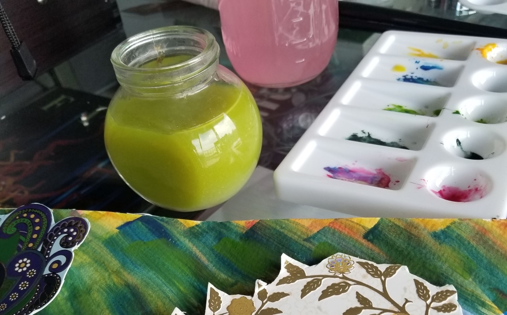 View of jars with colored water and a paint palette. In the foreground is a canvas with various colors and some paper cut outs.