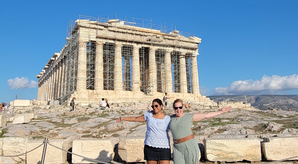 Two women standing in front of the Parthenon in Greece. The site has scaffolding around it.