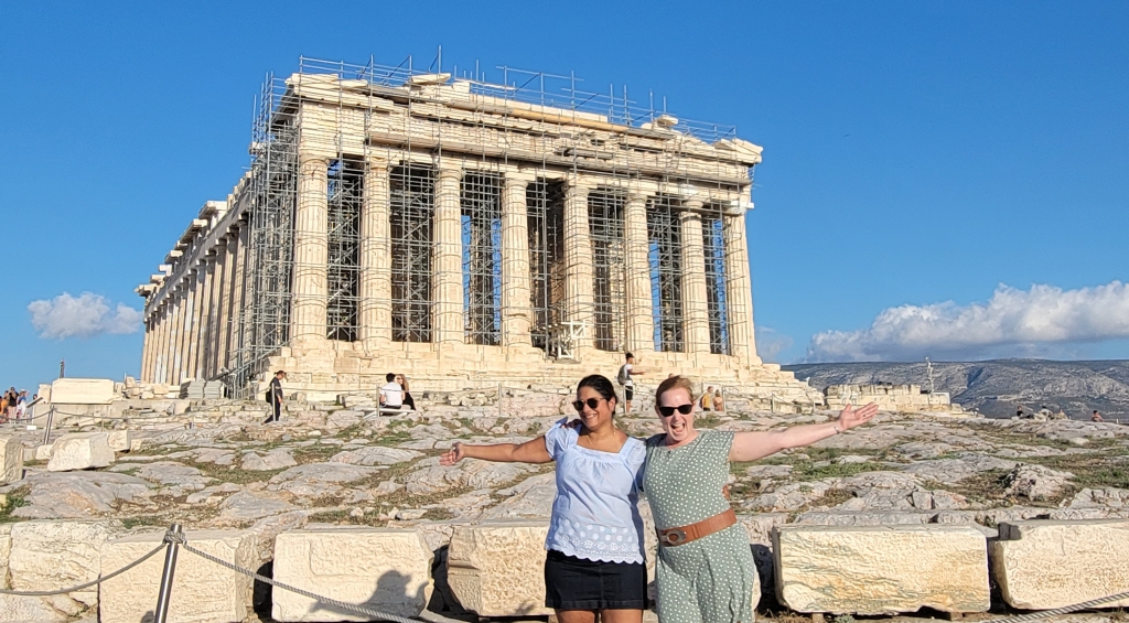 Two women standing in front of the Parthenon in Greece. The site has scaffolding around it.