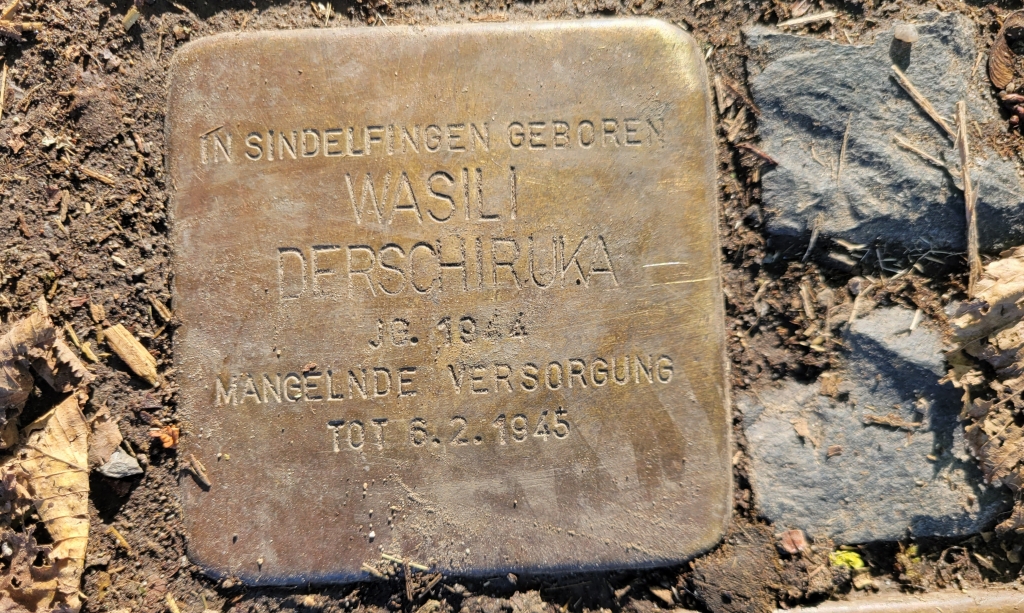 A square brass stone surrounded by grey stones. There is a name and inscription and a date of death engraved on the stone.