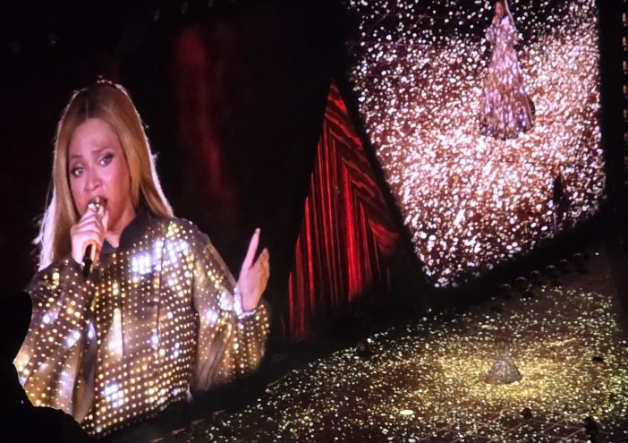 A woman on a screen is in a dress lit up in gold holding a microphone, next to it is another screen with a slightly wider view of the dress and the stage lighting up together while in the bottom right you can see the woman herself.