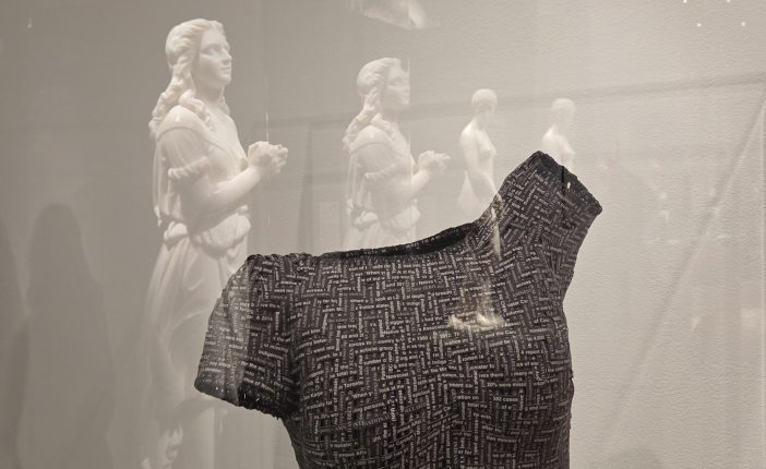 A view of the top part of a sculpture which is in the form of the top of a owman's torso. Reflected in the glass case are white marble sculptures of varying forms.